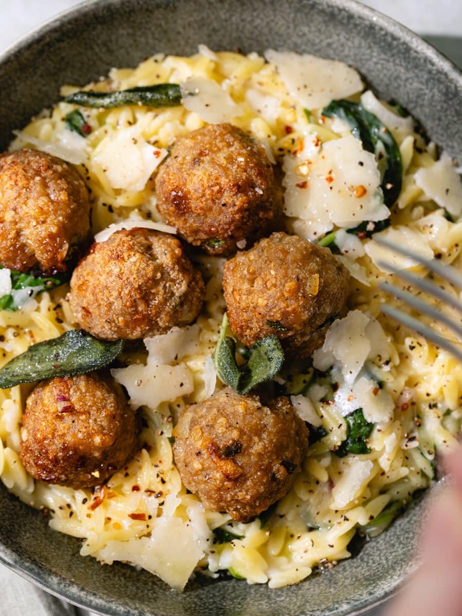 Gros plan sur des boulettes de poulet maison servies sur un orzotto crémeux au parmesan avec de la sauge croustillante et des copeaux de parmesan.