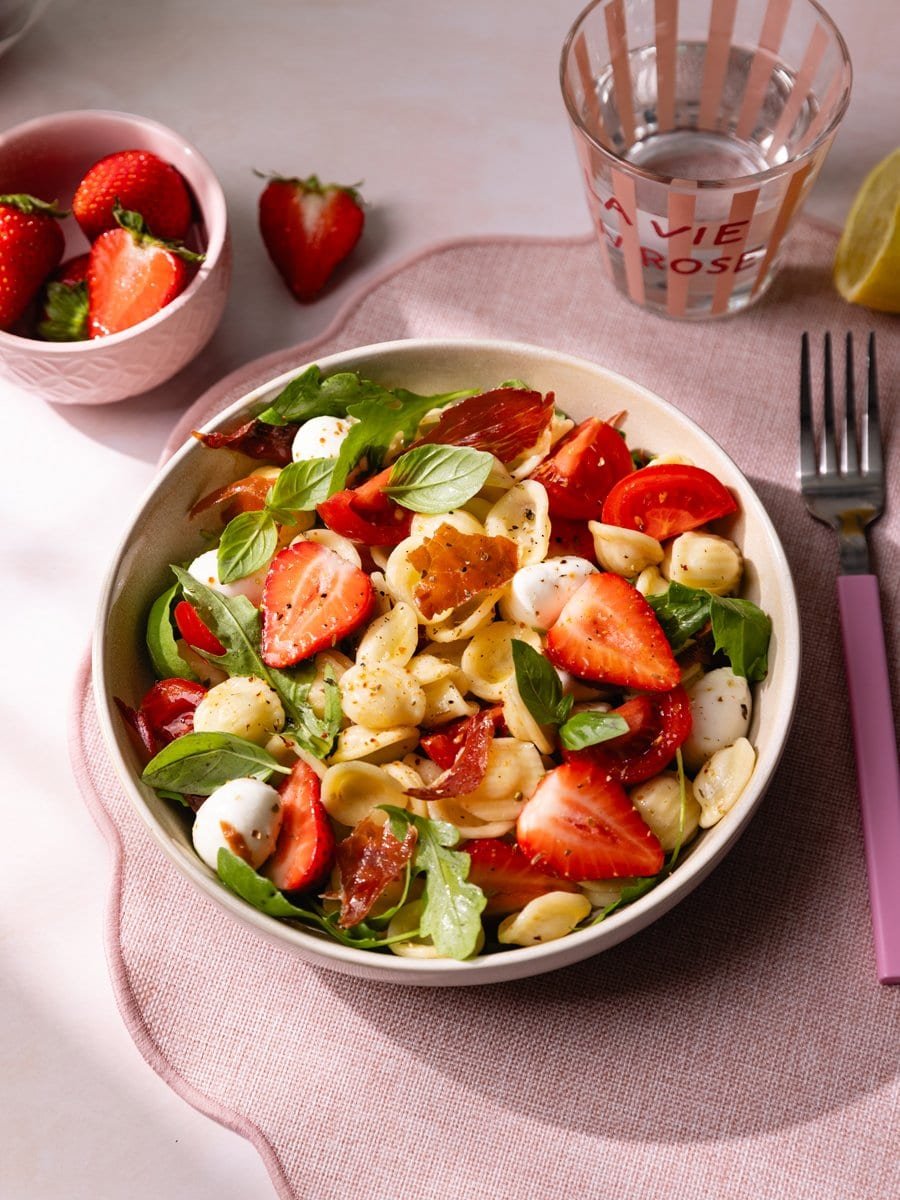 Bol de salade de pâtes aux fraises, mozzarella, tomates cerises, basilic et jambon cru servi sur une table estivale