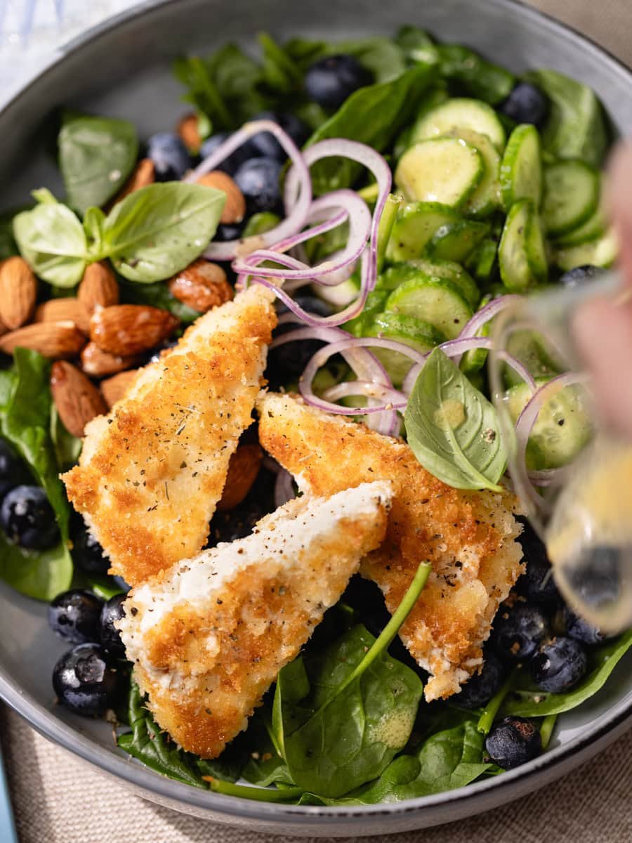 Salade fraîche aux épinards, myrtilles et feta panée croustillante servie avec concombre, oignon rouge et amandes dans un grand bol gris.