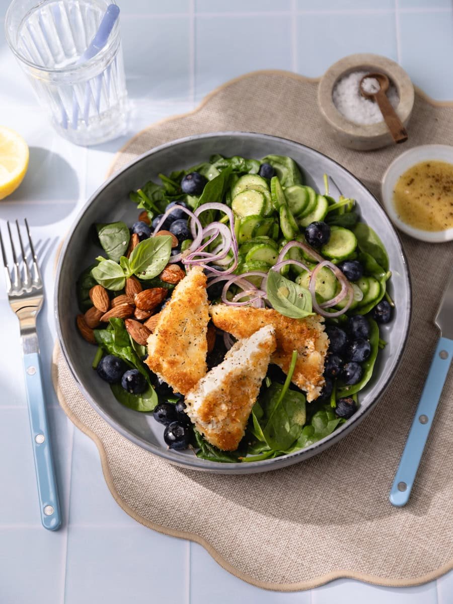 Salade fraîche aux épinards, myrtilles et feta panée croustillante servie avec concombre, oignon rouge et amandes dans un grand bol gris.