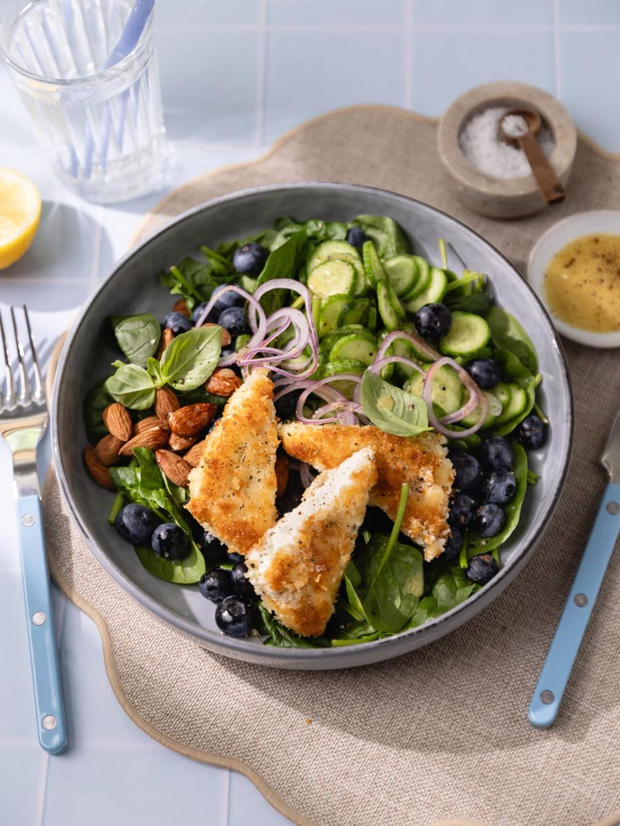 Salade fraîche aux épinards, myrtilles et feta panée croustillante servie avec concombre, oignon rouge et amandes dans un grand bol gris.