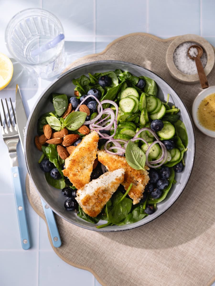 Salade fraîche aux épinards, myrtilles et feta panée croustillante servie avec concombre, oignon rouge et amandes dans un grand bol gris.