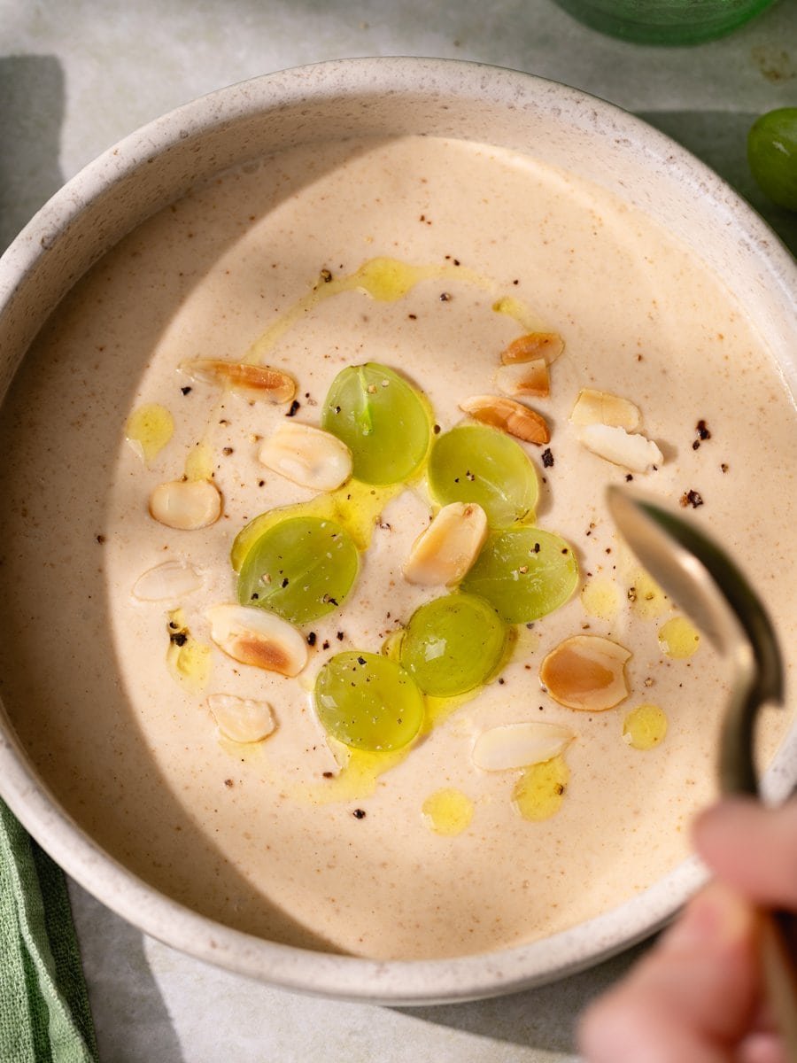 Cuillère s’approchant d’un ajo blanco crémeux décoré de raisins verts et d’amandes