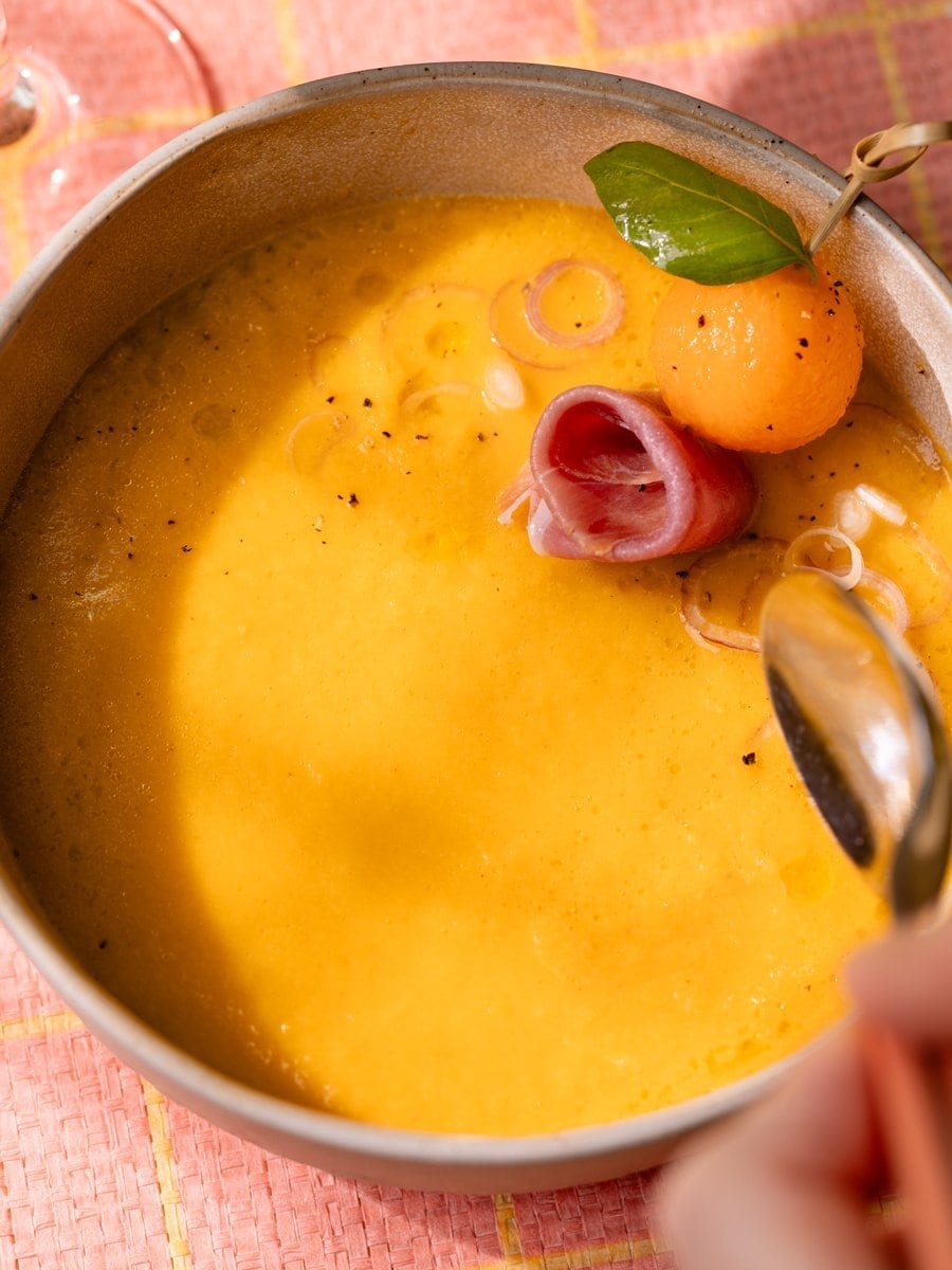 Cuillère plongeant dans un gaspacho au melon garni de jambon cru, de billes de melon et d'échalote