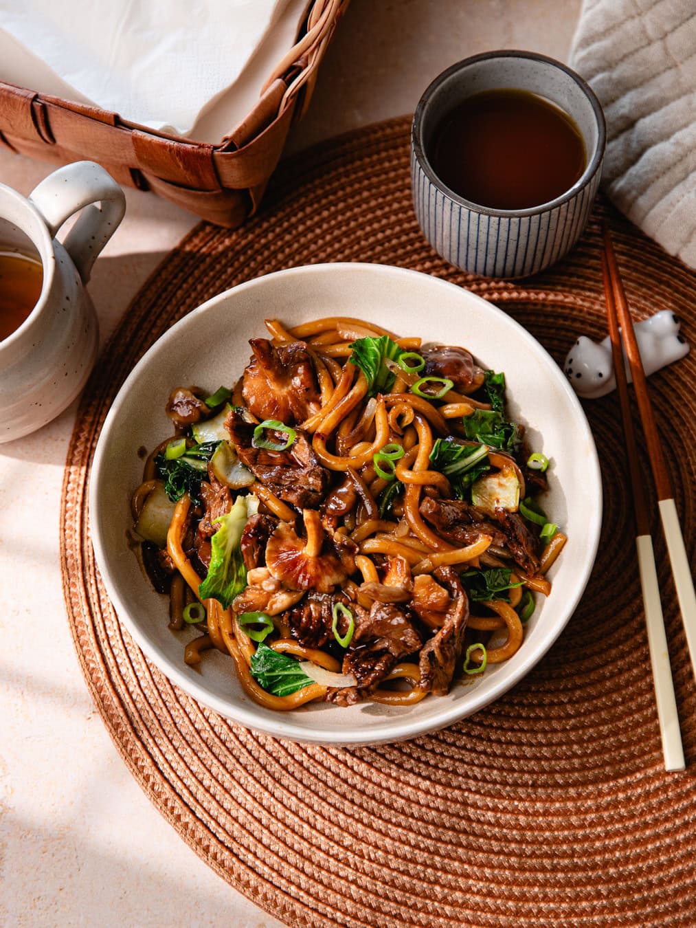 Un bol de nouilles udon sautées avec du bœuf, légumes et sauce savoureuse, servi avec une boisson chaude dans un cadre convivial.