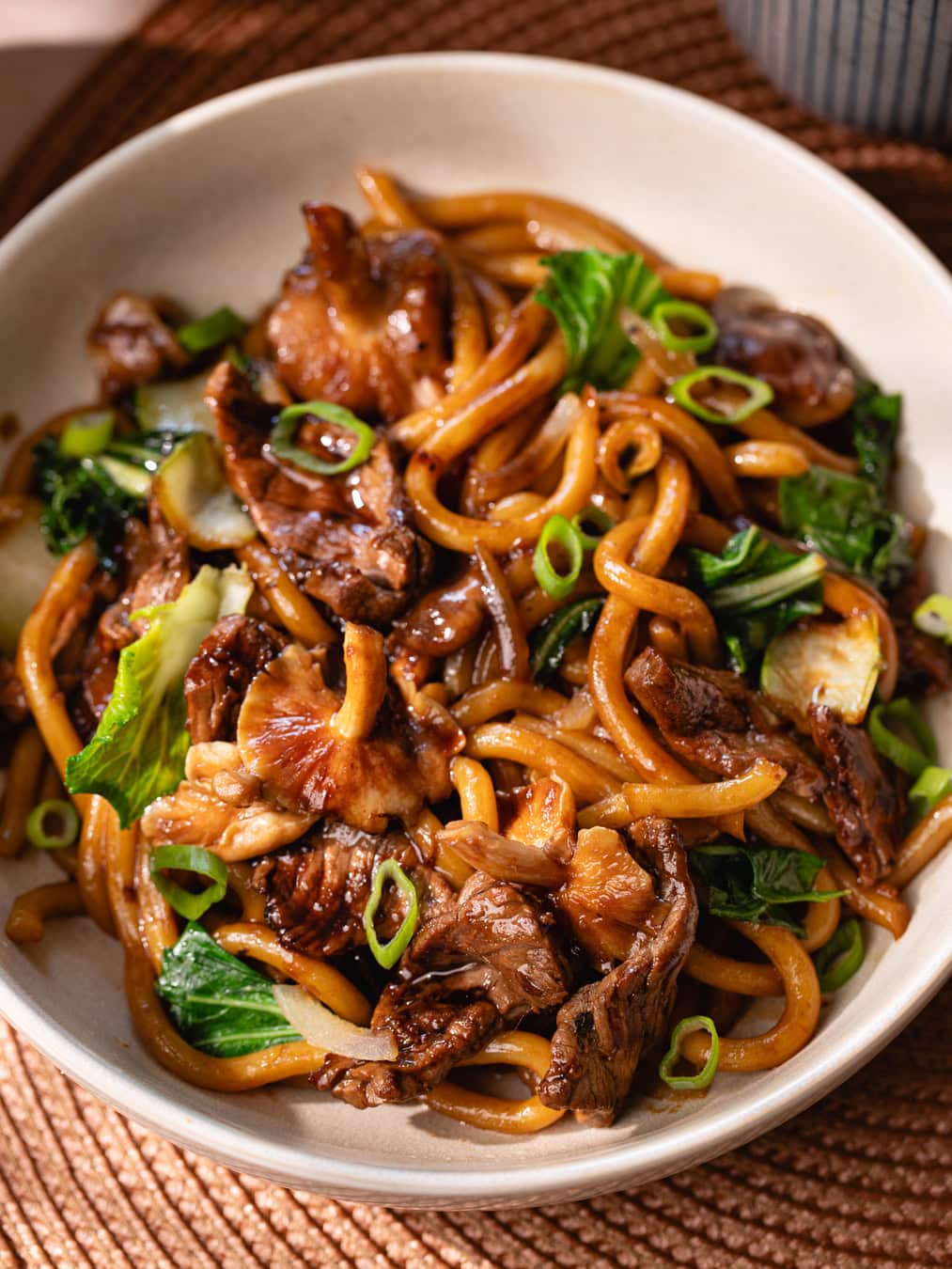 Un bol de nouilles udon sautées avec du bœuf, légumes et sauce savoureuse, servi avec une boisson chaude dans un cadre convivial.