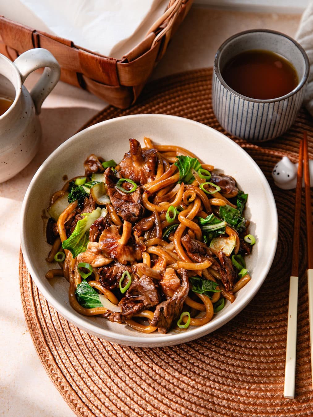 Un bol de nouilles udon sautées avec du bœuf, légumes et sauce savoureuse, servi avec une boisson chaude dans un cadre convivial.