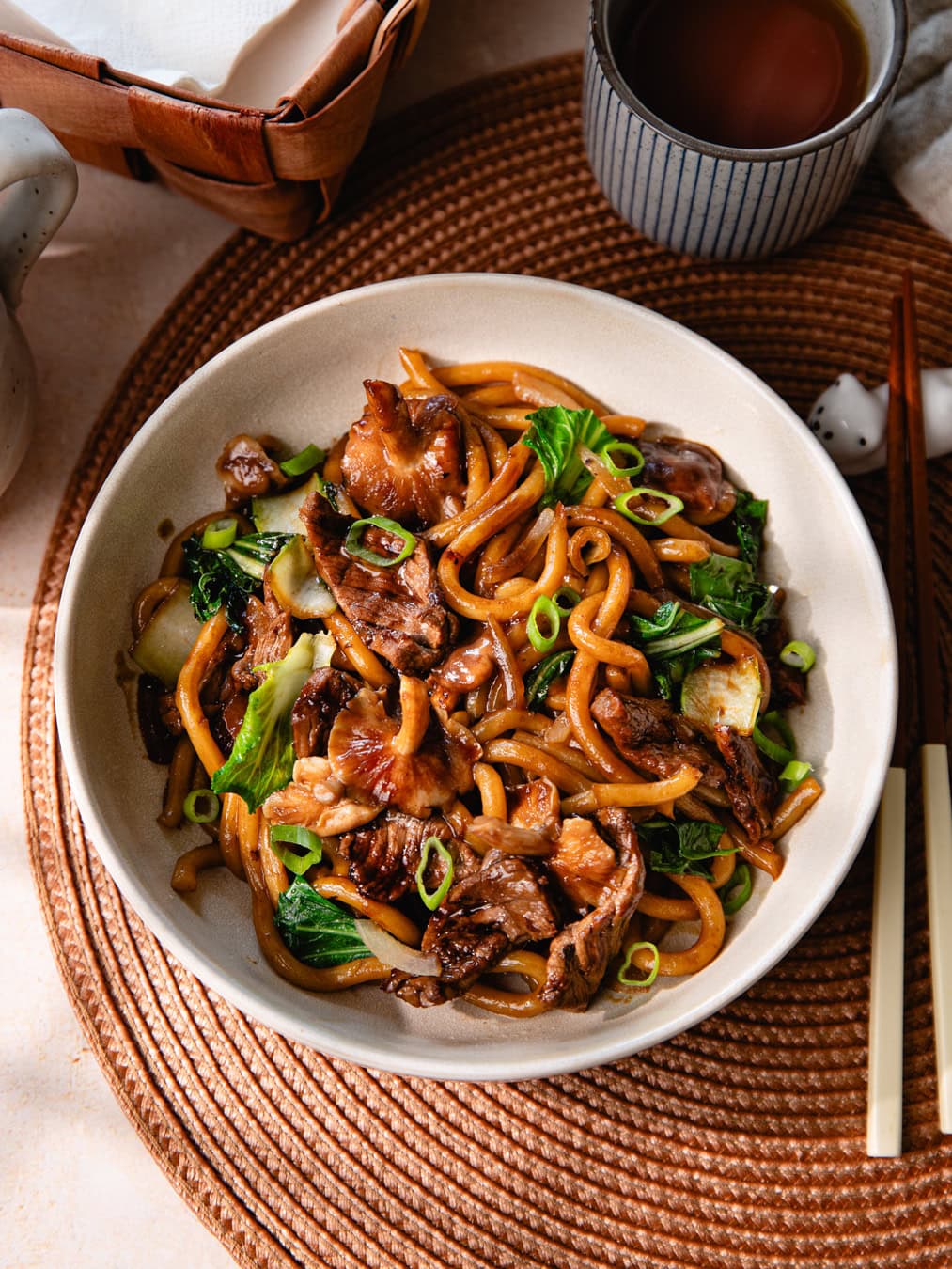 Un bol de nouilles udon sautées avec du bœuf, légumes et sauce savoureuse, servi avec une boisson chaude dans un cadre convivial.