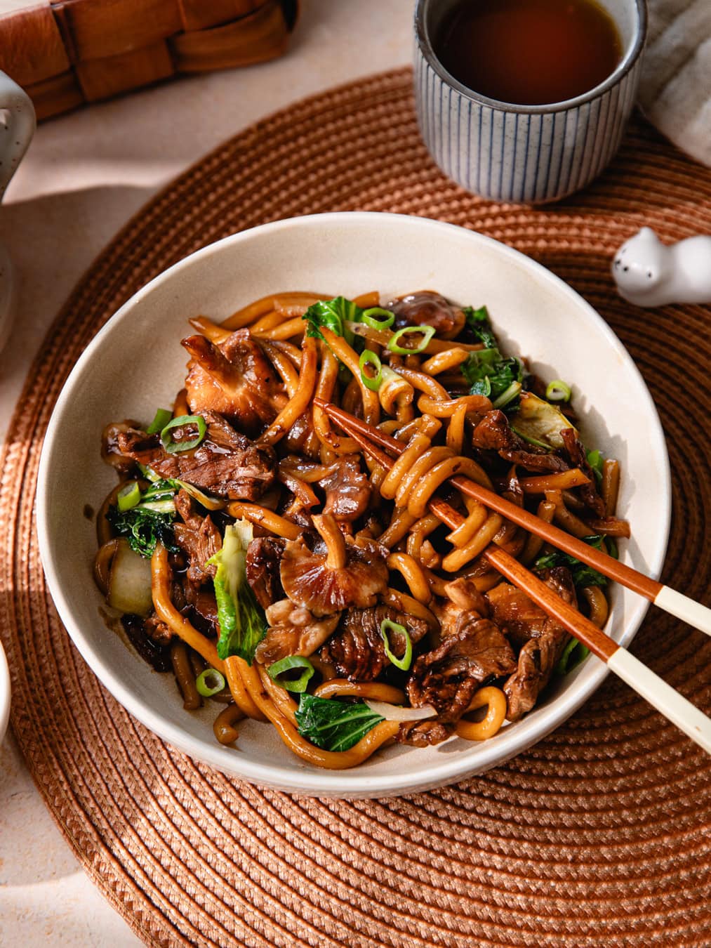Un bol de nouilles udon sautées avec du bœuf, légumes et sauce savoureuse, servi avec une boisson chaude dans un cadre convivial.