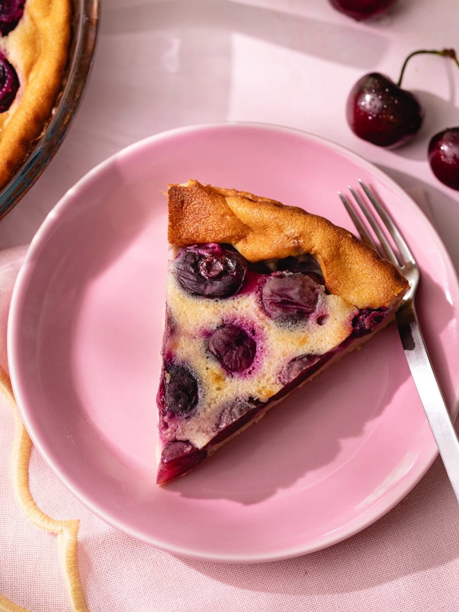 Clafoutis aux cerises maison servi sur une assiette rose avec part découpée et cerises fraîches en décoration