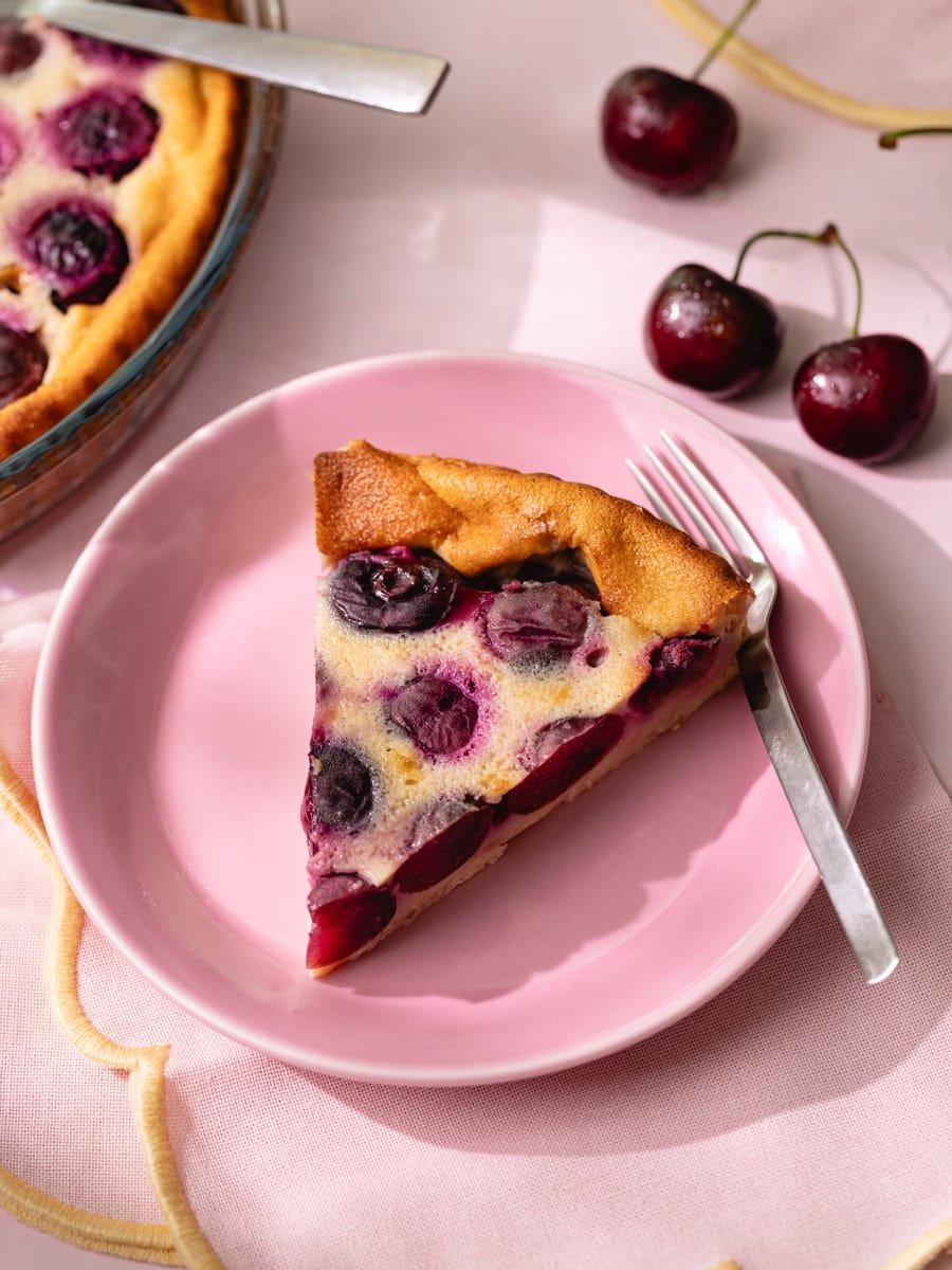 Clafoutis aux cerises maison servi sur une assiette rose avec part découpée et cerises fraîches en décoration