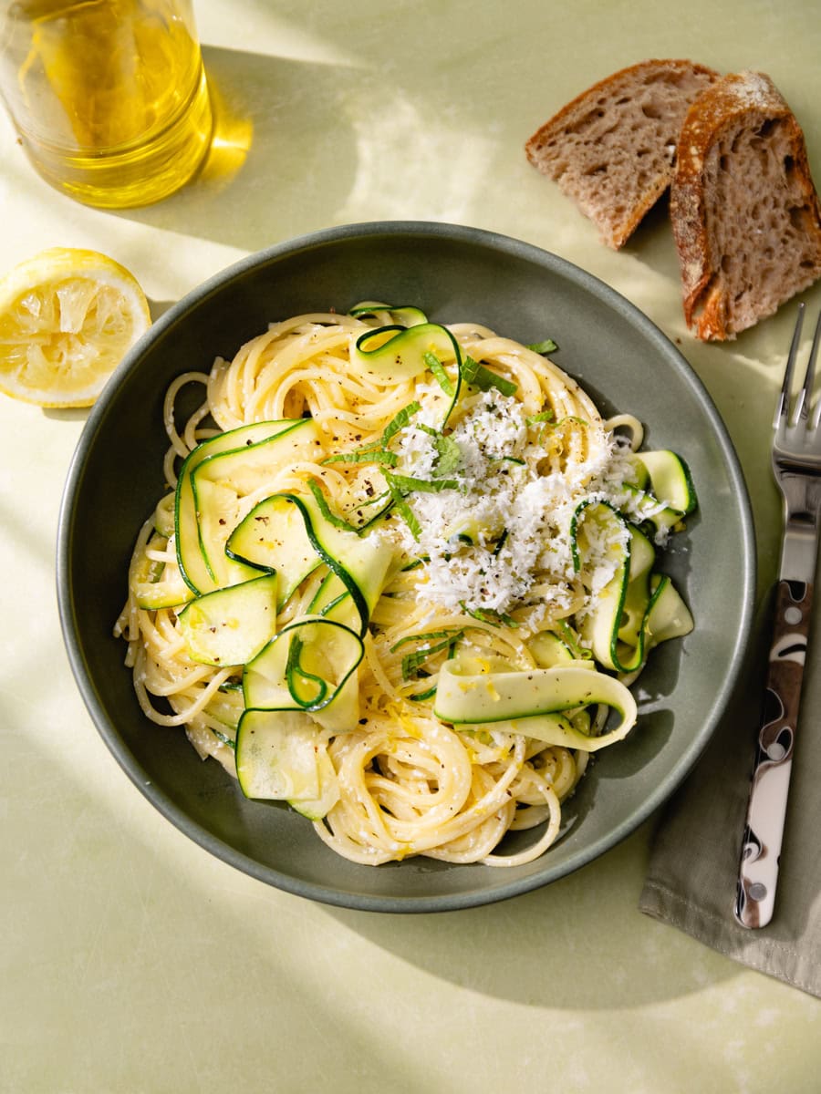 Assiette de spaghetti aux courgettes en tagliatelles, citron et ricotta salata râpée, servie avec menthe fraîche, huile d’olive et pain rustique sur une table claire ensoleillée.