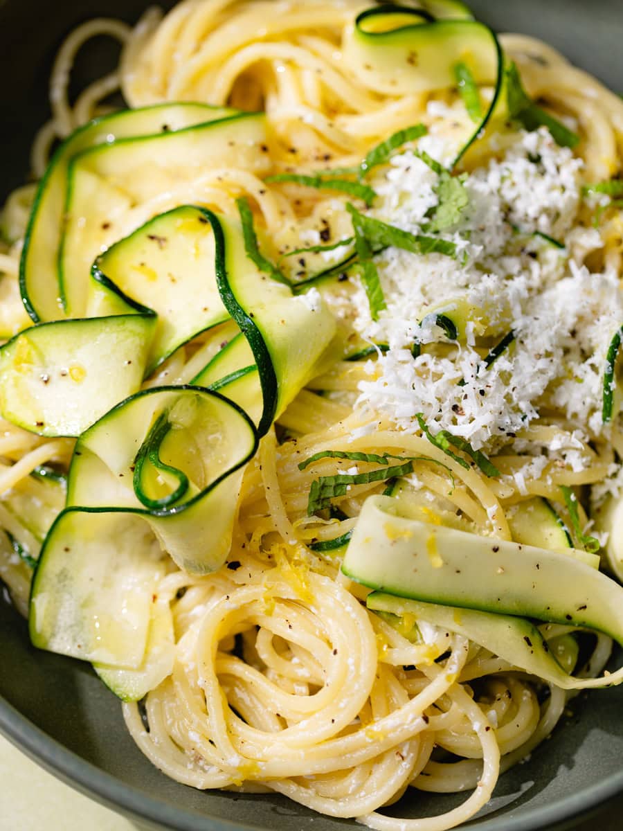 Assiette de spaghetti aux courgettes en tagliatelles, citron et ricotta salata râpée, servie avec menthe fraîche, huile d’olive et pain rustique sur une table claire ensoleillée.