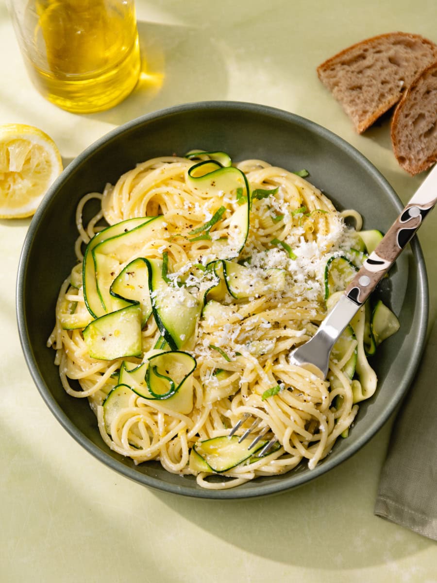 Assiette de spaghetti aux courgettes en tagliatelles, citron et ricotta salata râpée, servie avec menthe fraîche, huile d’olive et pain rustique sur une table claire ensoleillée.