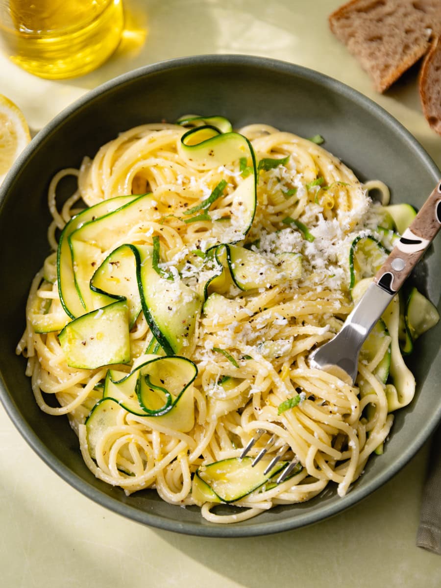Assiette de spaghetti aux courgettes en tagliatelles, citron et ricotta salata râpée, servie avec menthe fraîche, huile d’olive et pain rustique sur une table claire ensoleillée.