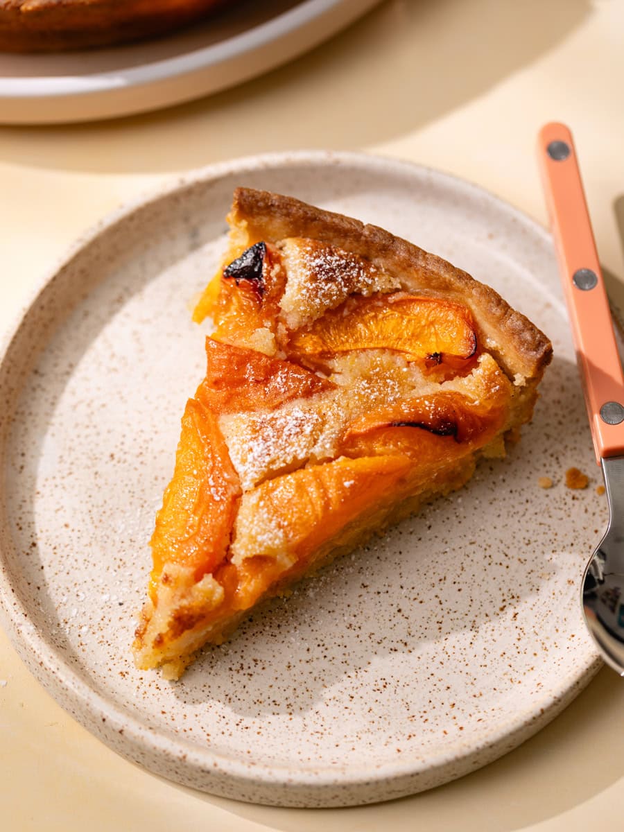 Part de tarte aux abricots et crème d’amande servie sur une assiette en grès, avec des abricots rôtis fondants et une pâte dorée légèrement saupoudrée de sucre glace.