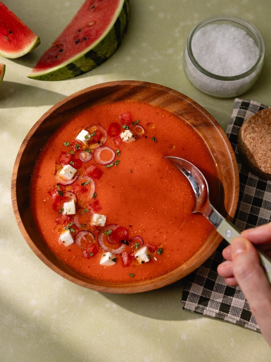 Main tenant une cuillère au-dessus d'un gaspacho pastèque tomate et feta