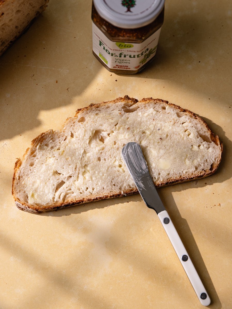 Grande tranche de pain blanc au levain recouverte de beurre avec couteau posé dessus vue du dessus